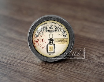 May include: A round, silver-colored cabinet knob with a glass insert. The glass insert features a vintage illustration of a bottle with a skull and crossbones, and the text "Laudanum - Poison". The surrounding text reads "Societe Cooperative Pharmacies Liegeoises Populaires".