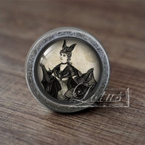 Könnte beinhalten: Ein silberner Knopf mit einem Glaseinsatz, der eine Vintage-Illustration einer Frau in einem Fledermauskostüm zeigt. Die Frau hält eine Fledermaus in der Hand.