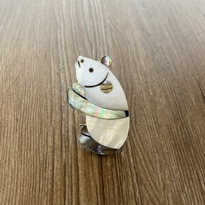 Puede incluir: Un broche de panda de madreperla blanca e iridiscente. El panda est&aacute; de pie sobre sus patas traseras con los brazos cruzados frente a &eacute;l.