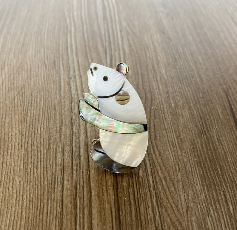 Puede incluir: Un broche de panda de madreperla blanca e iridiscente. El panda est&aacute; de pie sobre sus patas traseras con los brazos cruzados frente a &eacute;l.