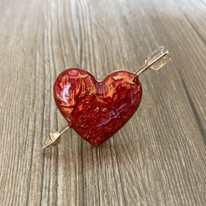 May include: A heart-shaped brooch with a gold arrow piercing through it. The heart is a vibrant red with a textured surface, and the arrow has a gold finish. The brooch is a romantic accessory.