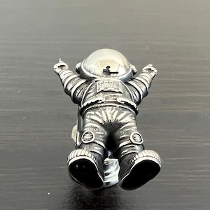 May include: A silver astronaut figurine with a textured surface and raised details. The astronaut is wearing a helmet and has its arms raised.