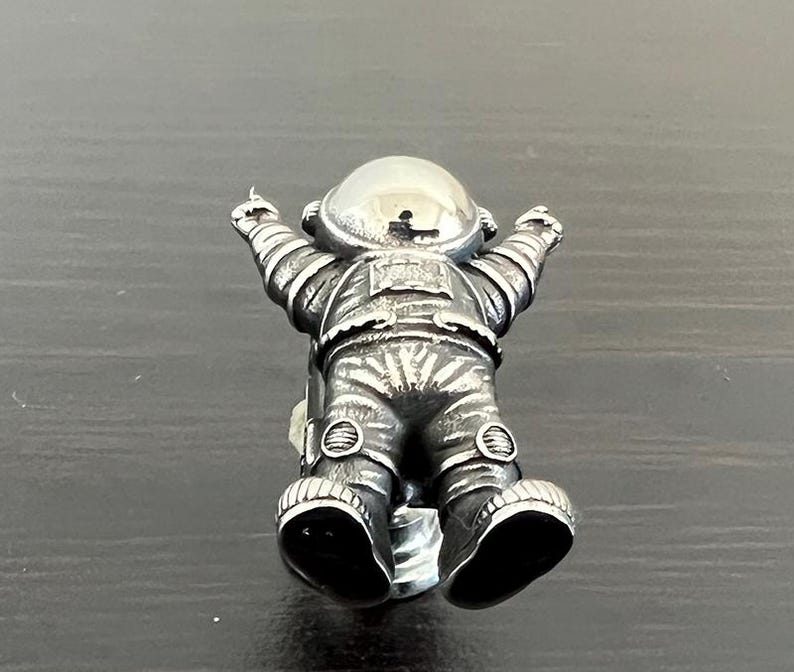 May include: A silver astronaut figurine with a textured surface and raised details. The astronaut is wearing a helmet and has its arms raised.