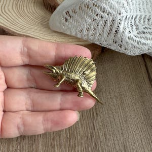 Op de afbeelding: Een gedetailleerd, antiek ogend messing Dimetrodon dinosaurusbeeldje. Het beeldje heeft een opvallende rugvin en ingewikkelde details, die een prehistorisch reptiel weergeven. De messing afwerking geeft het een vintage uitstraling, perfect voor verzamelaars.