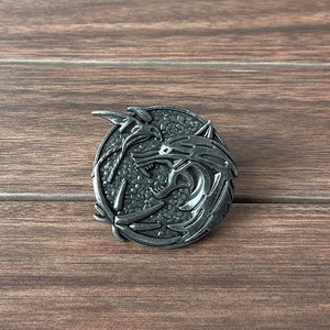 May include: A silver-toned metal pin featuring a stylized design of two wolves facing each other. The wolves are depicted in a circular shape with their heads and necks intertwined.