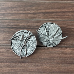 May include: Two silver-toned metal pins, one with a stylized bird design and the other with a stylized plant design.