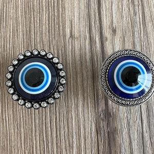 Può includere: Due pomelli per porte decorativi a forma di occhio turco con un design a occhio blu e bianco e una cornice in metallo argentato con un bordo di cristallo.