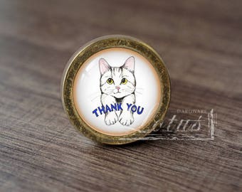 May include: A round, gold-coloured cabinet knob with a glass insert featuring a white cat with black stripes. The cat is looking at the viewer and has the words "Thank You" written in blue below it.