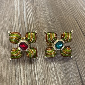 May include: Two gold-tone flower-shaped brooches with green and orange enamel. One brooch has a red gemstone in the centre, and the other has a green gemstone.