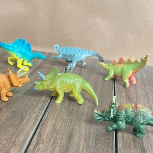 May include: A selection of six small dinosaur models in various colours. The dinosaurs are in shades of orange, green, blue, and grey. They are displayed on a wooden surface.