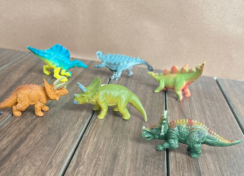 May include: A selection of six small dinosaur models in various colours. The dinosaurs are in shades of orange, green, blue, and grey. They are displayed on a wooden surface.