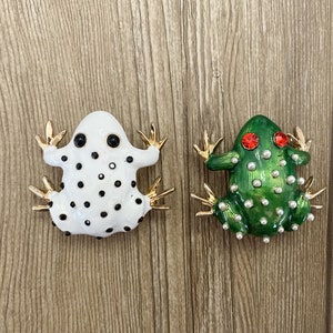 Może przedstawiać: Dwa broszki w kształcie żaby, jedna biała w czarne kropki i złote akcenty, druga zielona z białymi perełkowymi akcentami i czerwonymi oczami.