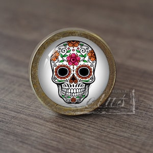 May include: A round, brass-coloured cabinet knob with a white, glass-like surface featuring a colourful sugar skull design. The skull is decorated with orange, pink, and green floral patterns.