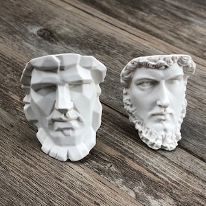 May include: Two white plaster sculptures of human faces. The face on the left is stylised and angular. The face on the right is more realistic and has a beard.