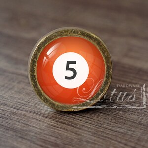 May include: A round, antique gold-colored cabinet knob with a red glass insert. The glass insert features a white circle with the number "5" in black.