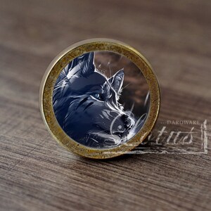 Può includere: Un pomello per armadio rotondo in ottone con un'immagine di testa di lupo al centro. Il lupo ha gli occhi azzurri e guarda a destra. L'immagine è circondata da un anello marrone scuro.