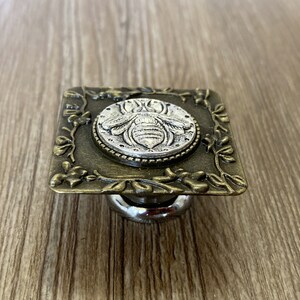 May include: A square-shaped, antique gold-toned metal cabinet knob with a silver bee design in the center. The knob has a decorative floral border.