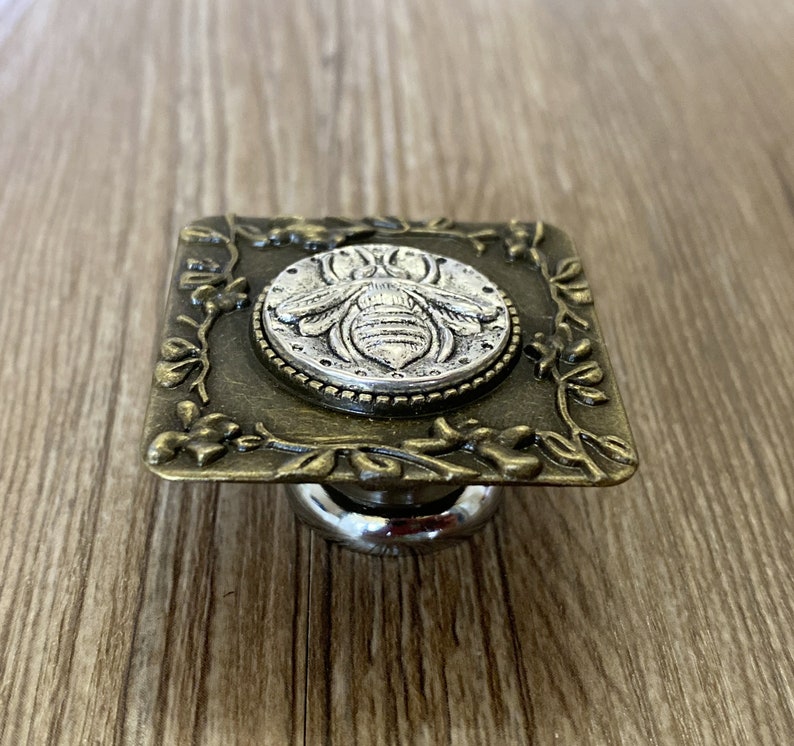 May include: A square-shaped, antique gold-toned metal cabinet knob with a silver bee design in the center. The knob has a decorative floral border.