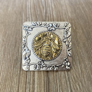 May include: A silver square ring with a gold bee design in the center. The bee is surrounded by a floral design.