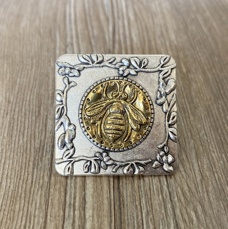 May include: A silver square ring with a gold bee design in the center. The bee is surrounded by a floral design.