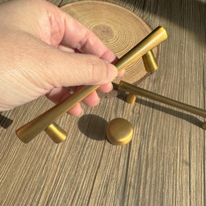 May include: A collection of brass cabinet hardware, including bar pulls and a round knob. The hardware has a warm, golden finish and is displayed on a wooden surface. The pulls are approximately 6 inches long.