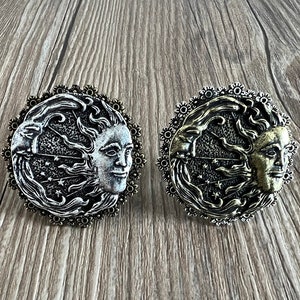May include: Two silver and gold tone metal brooches with a sun and moon design. The brooches are round and have a detailed design of a sun and moon with stars and clouds.