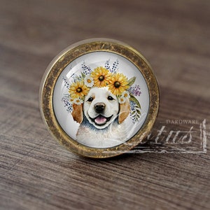 May include: Round cabinet knob with a brass finish. The knob features a watercolor image of a smiling golden retriever puppy wearing a crown of sunflowers and other flowers. The knob is set against a wooden surface.