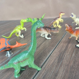 May include: A selection of plastic dinosaur toys in various colours, including green, brown, and orange. The toys include a Brachiosaurus, a Pterodactyl, and other dinosaur figures. They are arranged on a wooden surface.