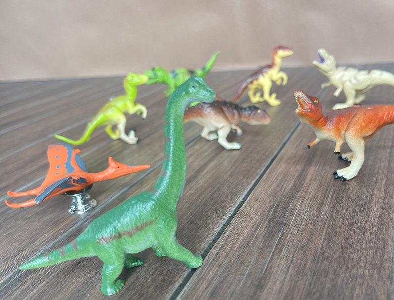 May include: A selection of plastic dinosaur toys in various colours, including green, brown, and orange. The toys include a Brachiosaurus, a Pterodactyl, and other dinosaur figures. They are arranged on a wooden surface.