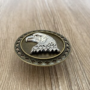 May include: A silver eagle head design on a round, bronze-coloured metal buckle with a decorative border.