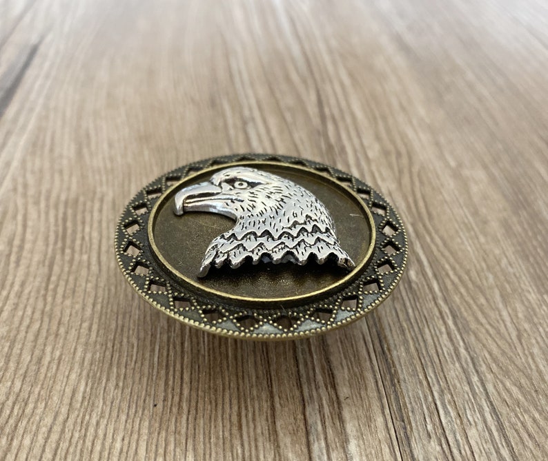 May include: A silver eagle head design on a round, bronze-coloured metal buckle with a decorative border.