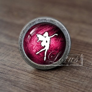 Può includere: Pomello rotondo per armadio con bordo in metallo color argento. Il pomello presenta un centro rosso intenso con una silhouette di fata bianca. La parola "Lotus" è visibile in basso a destra. Il pomello è su una superficie di legno.
