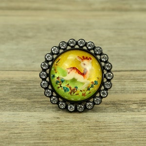 May include: A round, black, metal knob with a clear glass centre featuring a yellow background with a green and brown deer illustration. The knob is surrounded by a ring of clear rhinestones.