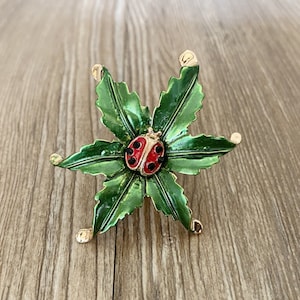 Könnte beinhalten: Eine goldfarbene Brosche mit einem grünen Emaille-Blumendesign und einem roten und schwarzen Marienkäfer in der Mitte.