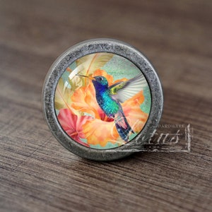 Puede incluir: Un pomo plateado con un inserto de vidrio que presenta un colibrí colorido posado sobre una flor naranja brillante con acentos rosados.