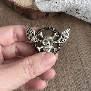 Puede incluir: Un emblema de calavera plateada con alas y huesos cruzados. El emblema es redondo con un fondo de color bronce y un borde con cuentas. La calavera tiene un diseño detallado, con las alas extendiéndose hacia afuera. El emblema se sostiene en una mano.