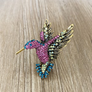 May include: A gold-toned hummingbird brooch with pink, blue, and yellow rhinestones. The hummingbird is in flight with its wings spread out.