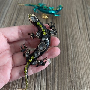 May include: A black and green jeweled lizard brooch with clear rhinestones. The lizard is posed with its legs outstretched and its tail curled.