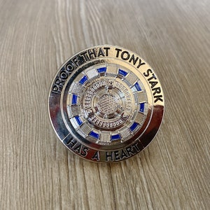 Peut inclure: Épinglette en métal argenté avec un motif bleu et blanc au centre. L'épingle porte l'inscription "Proof that Tony Stark has a heart".