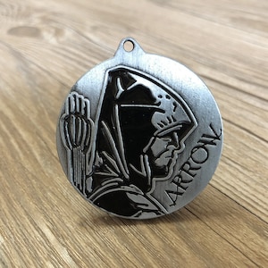 May include: A silver metal keychain with a black silhouette of a hooded figure holding an arrow. The word "ARROW" is written vertically on the keychain.
