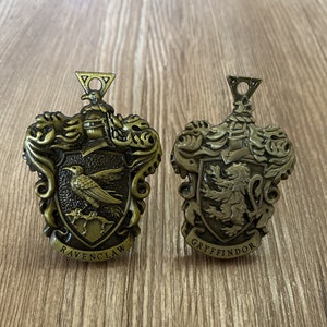 Peut inclure: Deux blasons de maison Harry Potter en métal doré. Le blason de gauche est pour Serdaigle et représente un corbeau avec une couronne. Le blason de droite est pour Gryffondor et représente un lion avec une couronne.