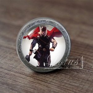 Könnte beinhalten: Ein runder, silberfarbener Schrank-Knopf mit einem Glaseinsatz, der eine Comic-Illustration von Thor zeigt, einem Superhelden mit einem roten Umhang und einem Hammer.