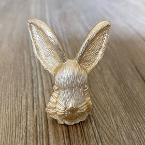 May include: A gold-colored bunny head figurine with detailed fur texture. The bunny has long ears and a detailed face. The figurine is made of metal and is likely a decorative item. The bunny is a symbol of spring and Easter.