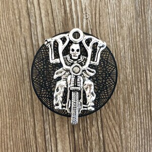 Op de afbeelding: Een zilverkleurige hanger met een skelet dat een motorfiets rijdt. De hanger is rond en heeft een zwarte achtergrond met een decoratief patroon.