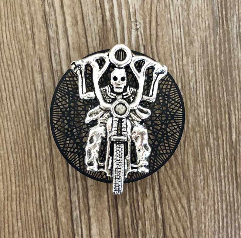 Op de afbeelding: Een zilverkleurige hanger met een skelet dat een motorfiets rijdt. De hanger is rond en heeft een zwarte achtergrond met een decoratief patroon.