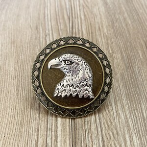 May include: A silver eagle head with a detailed design is featured on a round, bronze-coloured pendant with a geometric border.