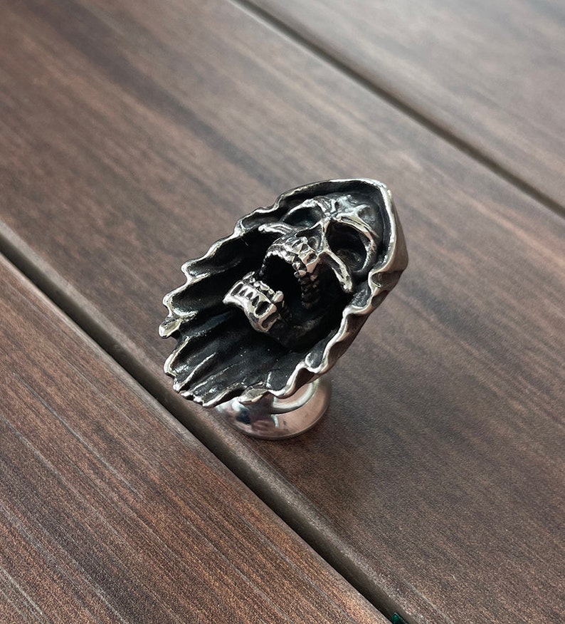 Op de afbeelding: Een zilverkleurige kastknop in de vorm van een schreeuwende schedel, met gedetailleerde tanden en een gedrapeerde sluier. De knop is gemonteerd op een ronde basis. Deze decoratieve hardware is ontworpen voor meubels en andere decoratieve doeleinden.