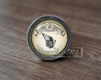 May include: A round, antique-style metal knob with a cream-colored label featuring a black and white wing illustration. The label includes French text and is set against a dark wooden surface.