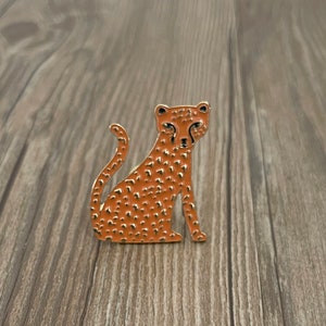 Peut inclure: Une épingle en métal dorée avec un motif de guépard orange. Le guépard est assis, la queue enroulée derrière lui.