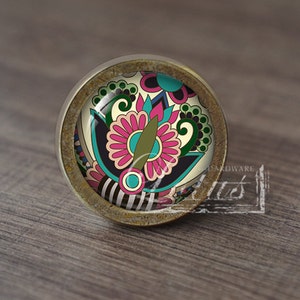 May include: A round, brass cabinet knob with a colourful, floral design. The knob has a distressed finish and a clear glass insert.